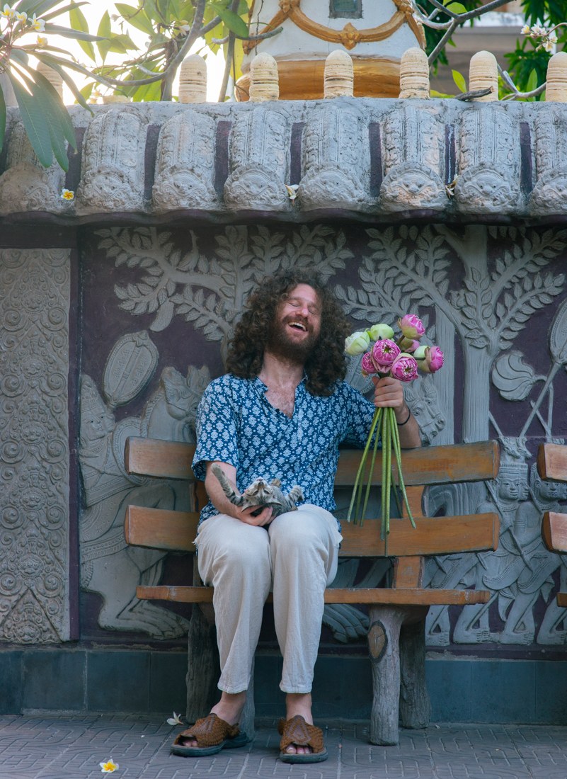 Director Jake Wachtel laughing on a temple bench in Cambodia, holding lotus flowers and a kitten